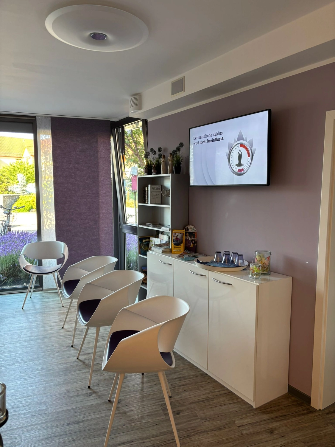 Frauenarztpraxis Dr. A. Dietrich - Gesundheitsdienste für Frauen - Ein Wartezimmer in einer Klinik mit weißen Stühlen, einem Aufbewahrungsschrank und einem an der Wand montierten Fernseher.