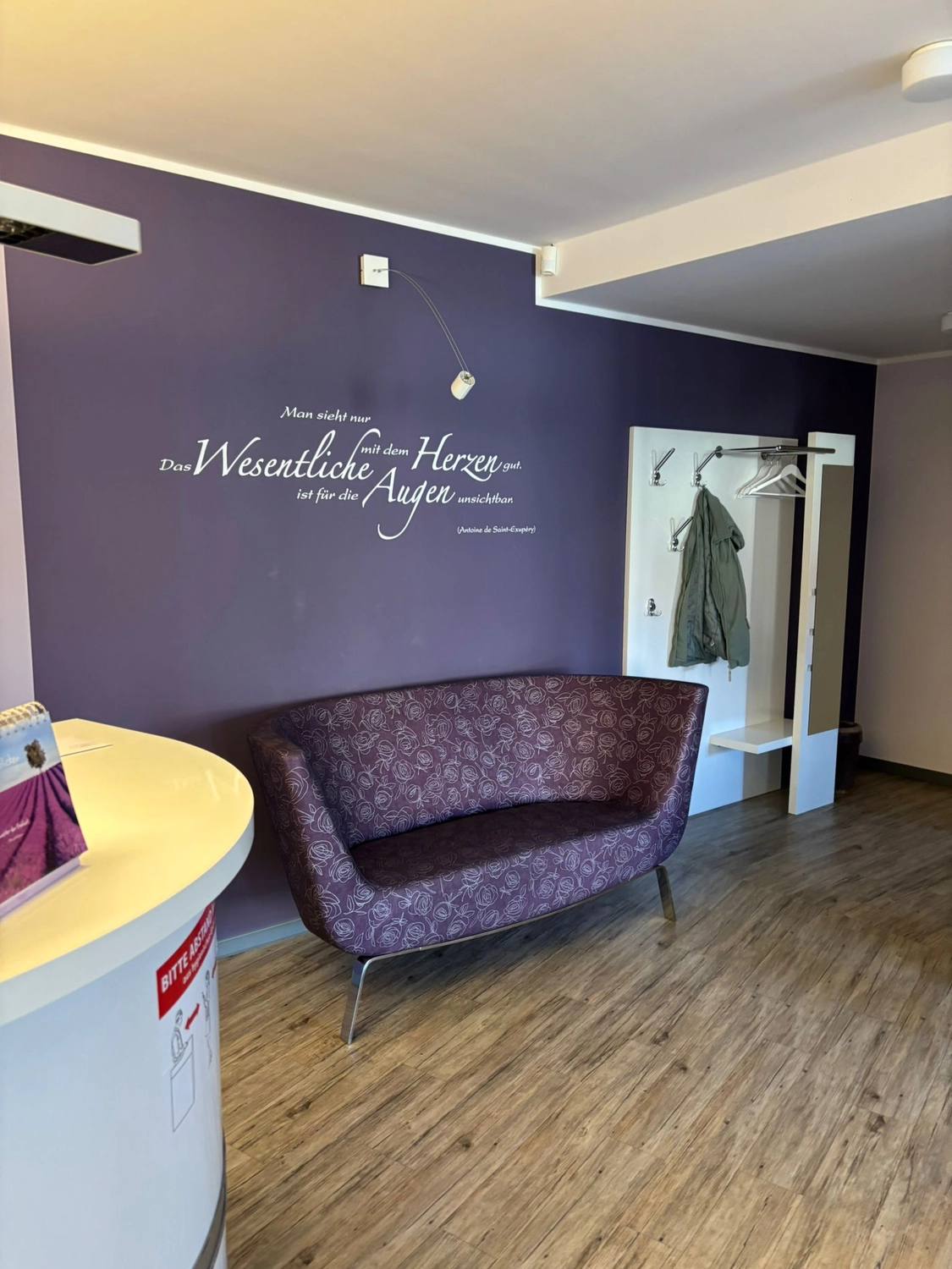 Frauenarztpraxis Dr. A. Dietrich - Gesundheitsdienste für Frauen - Ein in Lila gehaltener Wartebereich mit einem gemusterten Sofa vor einer dunkelvioletten Wand mit weißer Schrift. Links ist ein kleiner weißer Empfangstresen zu sehen, rechts im Hintergrund eine weiße Garderobe.