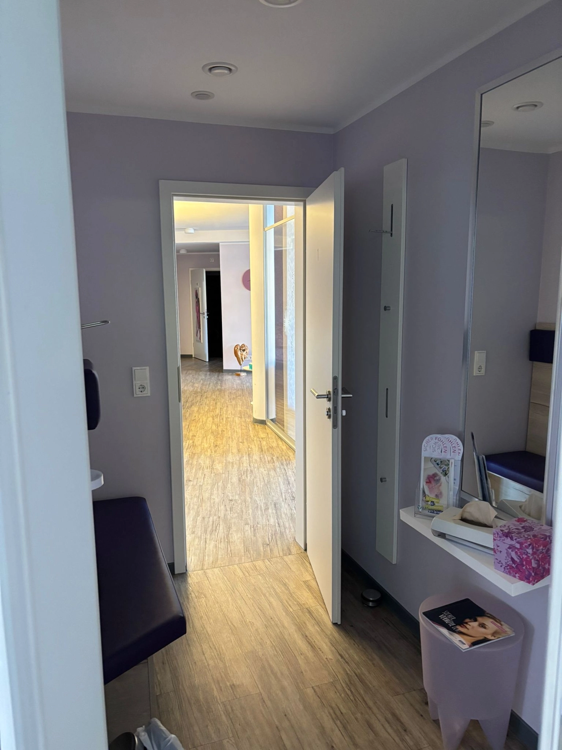 Frauenarztpraxis Dr. A. Dietrich - Gesundheitsdienste für Frauen - Ein hell erleuchteter, moderner Flur mit hellem Holzboden, einer offenen weißen Tür und einem kleinen Raum mit Spiegel.