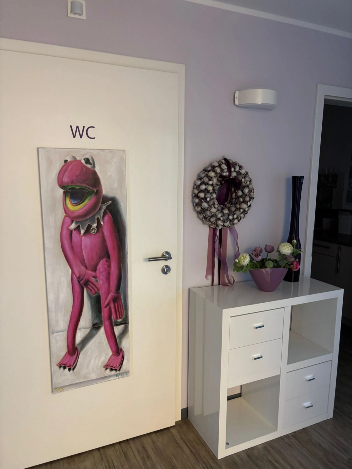 Frauenarztpraxis Dr. A. Dietrich - Gesundheitsdienste für Frauen - Eine weiße Tür mit der Aufschrift „WC“ zeigt ein vertikales Poster einer pinkfarbenen, Kermit-ähnlichen Figur. Daneben steht ein kleiner weißer Schrank mit Dekoration, darunter ein Blumenkranz und eine Blumenvase.