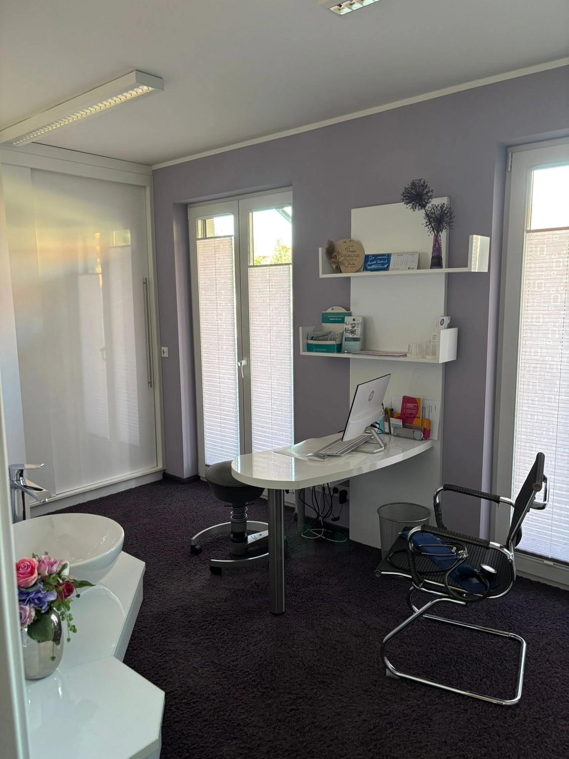 Frauenarztpraxis Dr. A. Dietrich - Gesundheitsdienste für Frauen - Ein Büro oder ein Untersuchungsraum mit einem geschwungenen weißen Schreibtisch, einem modernen schwarzen Freischwingerstuhl und integrierten Regalen vor einer lavendelfarbenen Wand.